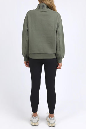 A person faces away from the camera, wearing a loose green Foxwood College Zip Crew with an embroidered collar, black leggings, and white sneakers against a plain white background.