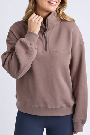 A woman wears the Foxwood College Zip Crew, a loose mauve cotton pullover, with dark fitted leggings. Her hand rests near the collar, her wavy hair is visible and her face is partially out of frame—ideal for casual outings.