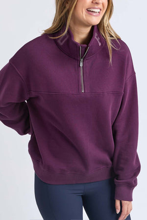 A woman smiles with one hand on her hip, wearing the Foxwood College Zip Crew—a purple pullover with an embroidered collar, high quarter-zip, and made from 100% soft cotton—set against a plain light background.