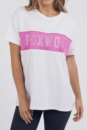 The woman models the Foxwood Connection Tee, a white 100% cotton T-shirt featuring a pink stripe and bold "FOXWOOD" text, paired with black pants against a simple backdrop, her face unseen.