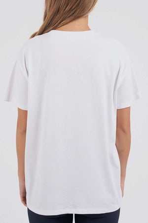 Seen from the back, a person with long, light brown hair models the Foxwood Connection Tee—a plain white T-shirt made of 100% cotton—paired with dark pants. They stand against a neutral background, embodying effortless leisurewear style.