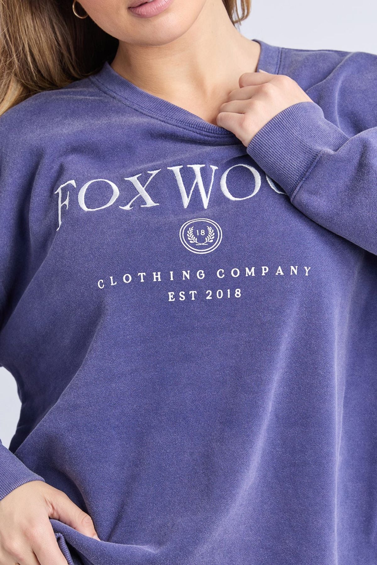 A woman wearing the Foxwood Core Crew, a loose-fitting faded purple cotton sweatshirt with "FOXWOOD CLOTHING COMPANY EST 2018" in white letters, holds the neckline with one hand.