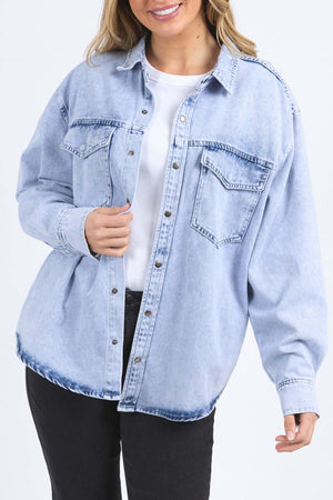 A woman smiles in the Foxwood Daisy Over Shirt, a relaxed fit light blue denim button-up made from 100% cotton, styled over a white top and black pants with one hand on the shirt, against a plain light background.
