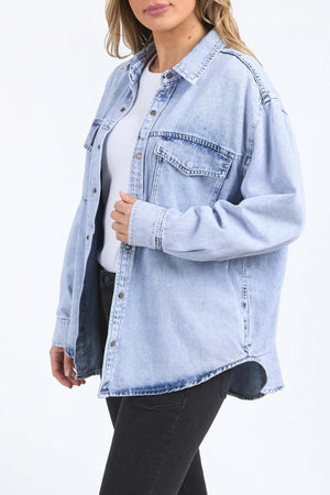 Wearing the Foxwood Daisy Over Shirt—light blue, 100% cotton, oversized with long sleeves and front pockets—over a white top and black jeans, a person stands against a plain white background.