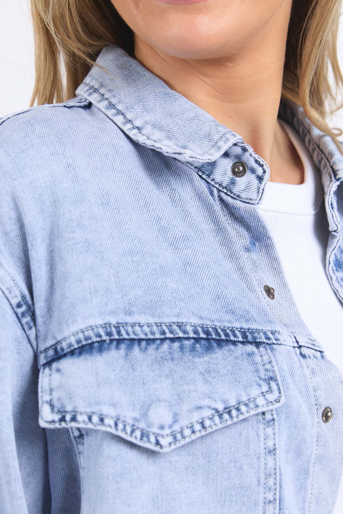 Close-up of a person wearing the Foxwood Daisy Over Shirt, a relaxed fit light blue denim jacket made from 100% cotton, layered over a white shirt. Focus is on the collar, upper chest, and shoulder; part of the face is partially shown.