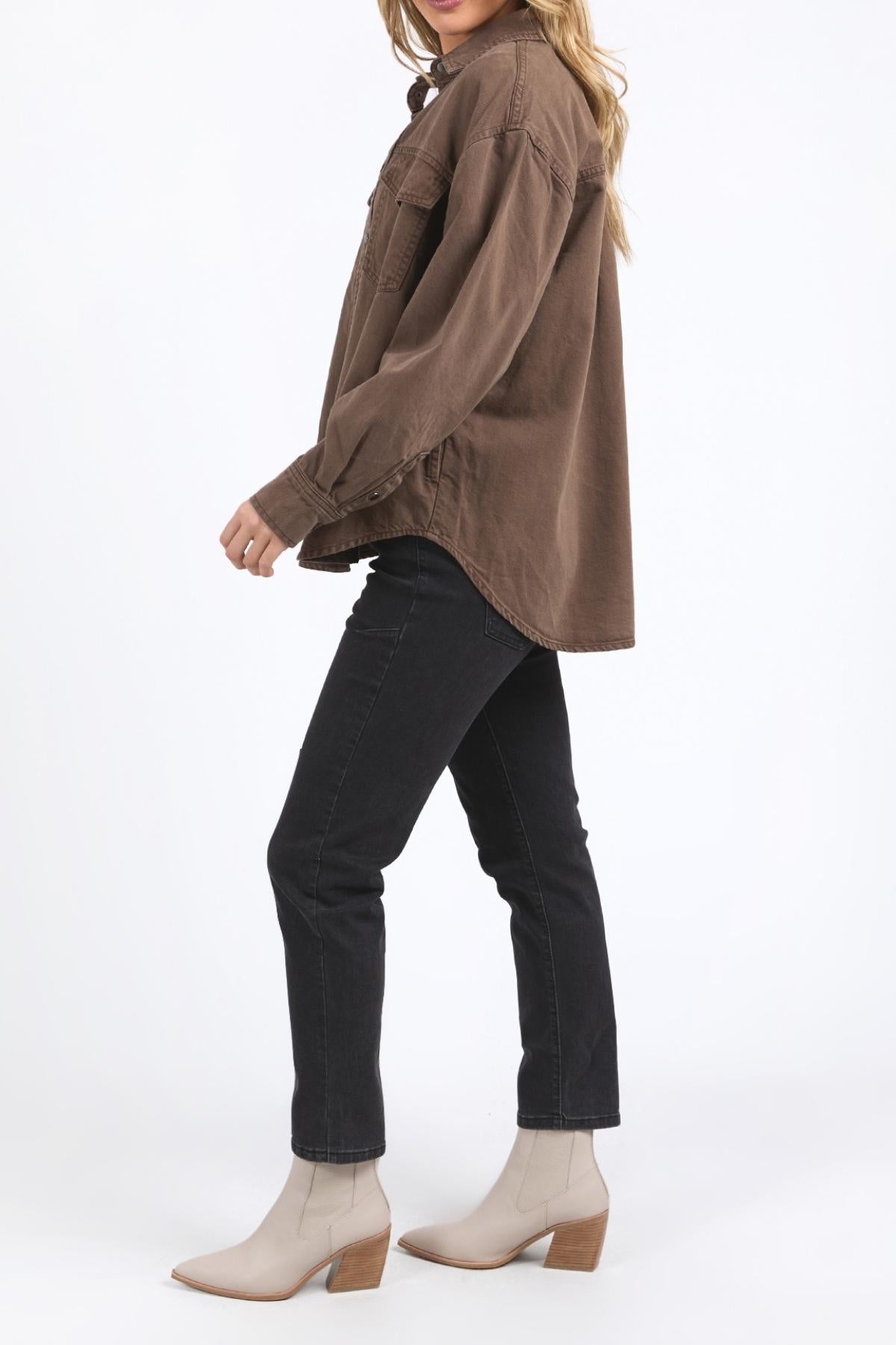 A person stands sideways against a plain white background, wearing the Foxwood Daisy Overshirt in brown cotton with a button-through placket, paired with black jeans and beige ankle boots.