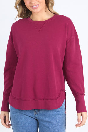 Woman wearing a maroon Foxwood Delilah Crew jumper with long sleeves and raw-edge details, paired with blue jeans. She smiles against a plain light background, her face partially visible.
