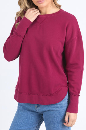 A woman wears the Foxwood Delilah Crew, a long-sleeved, dark pink jumper with a crew neck, paired with blue jeans. She stands on a plain white background with one hand touching her hair.