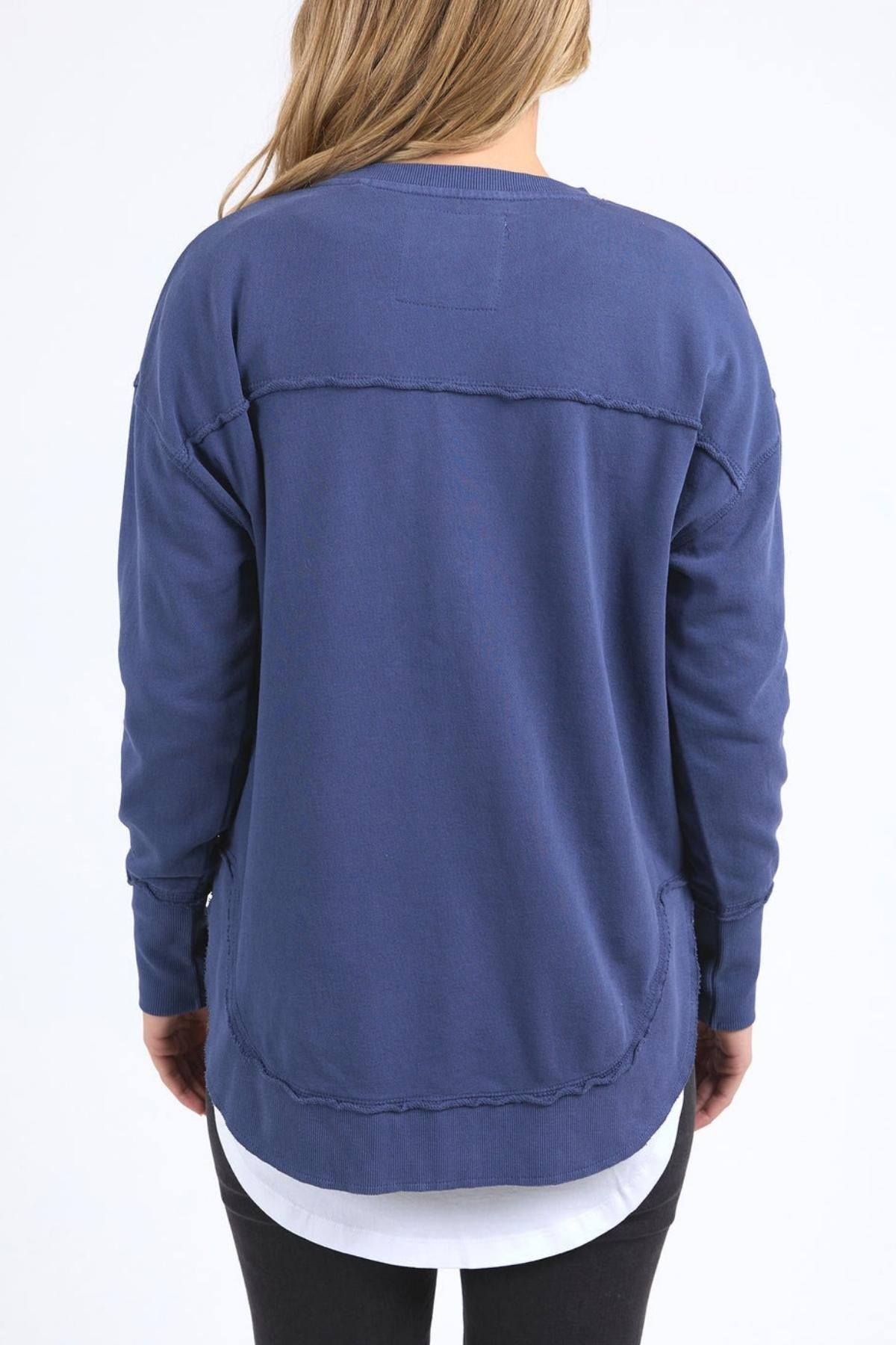 A person with long, light brown hair is shown from the back wearing the Foxwood Delilah Crew in blue over a white shirt and black pants, standing against a plain white background.