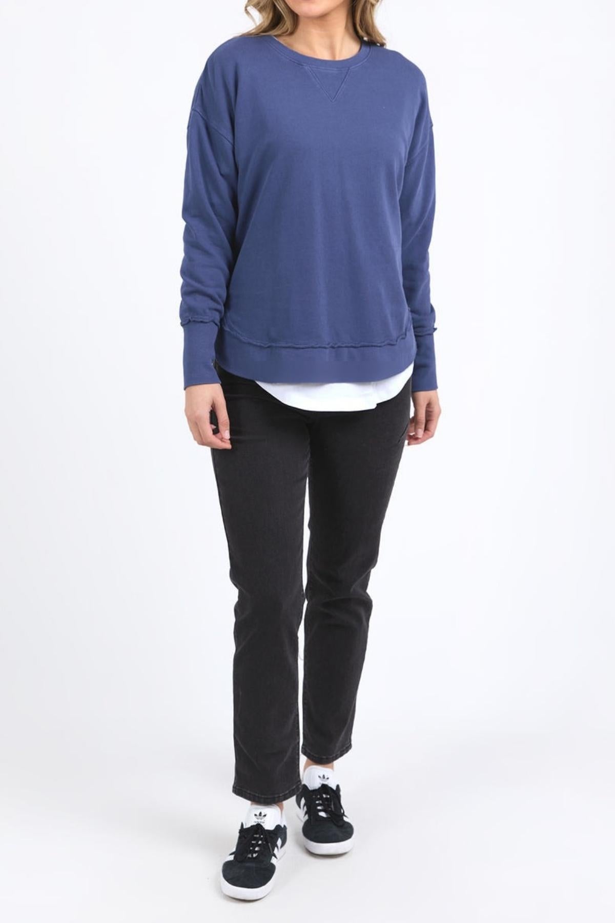 A woman stands against a white background wearing the Foxwood Delilah Crew in blue over a white shirt, paired with black jeans and black sneakers with white soles. Her hands are by her sides, and her face is not shown.