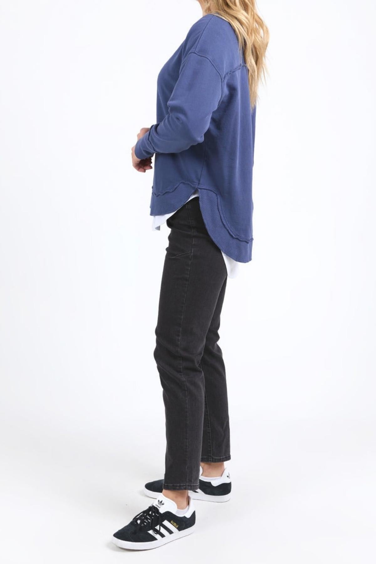 A woman stands sideways wearing the blue Foxwood Delilah Crew, black jeans, and black Adidas sneakers with white soles. Her long blonde hair is down against a plain white background.