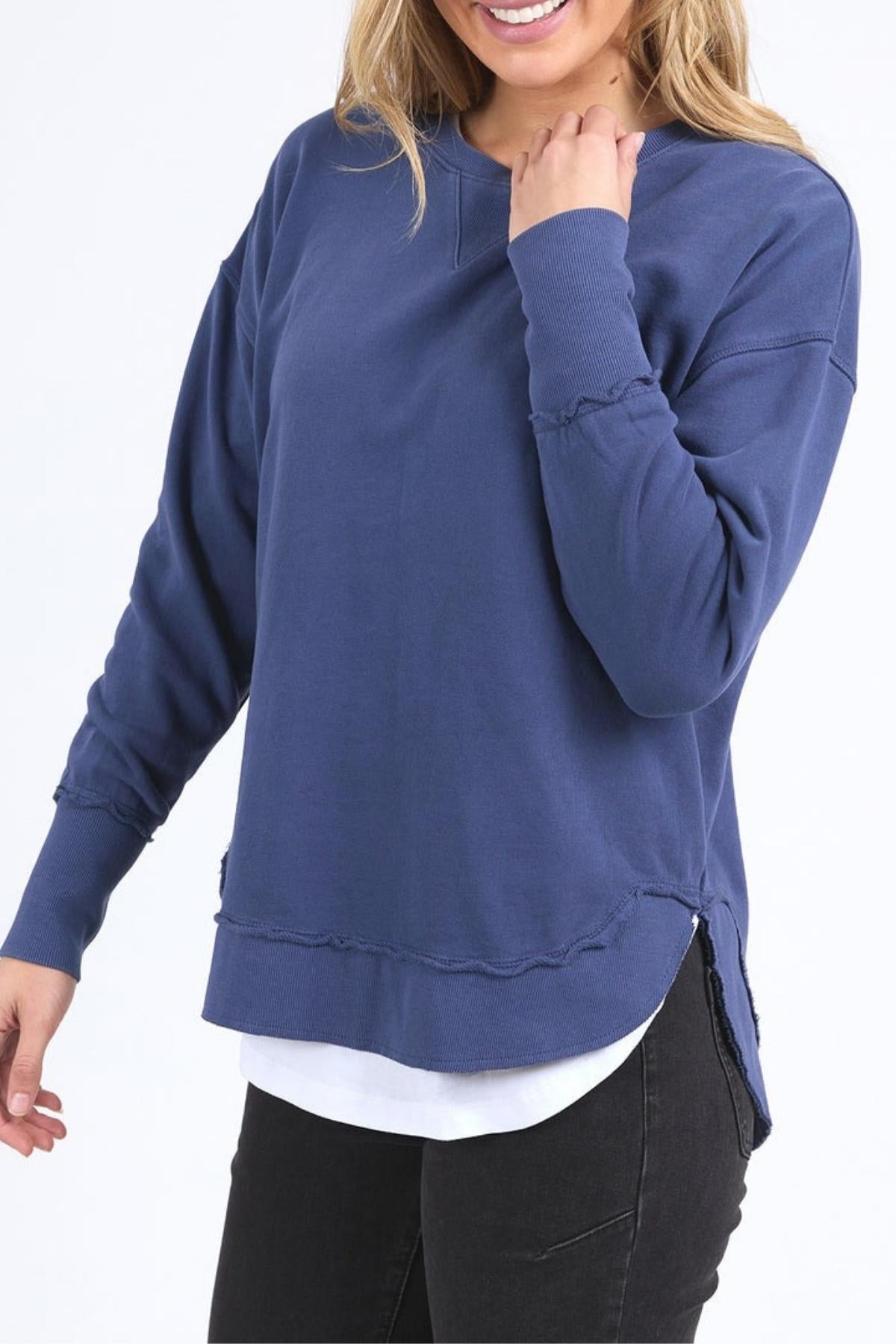 A person wearing the Foxwood Delilah Crew in blue over a white shirt with black jeans, smiling and posing against a plain white background.