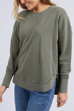 A person wears the Foxwood Delilah Crew, a loose cotton crew neck sweatshirt in olive green with raw-edge hems, paired with blue jeans. They smile with their head slightly turned and their left arm relaxed at their side.