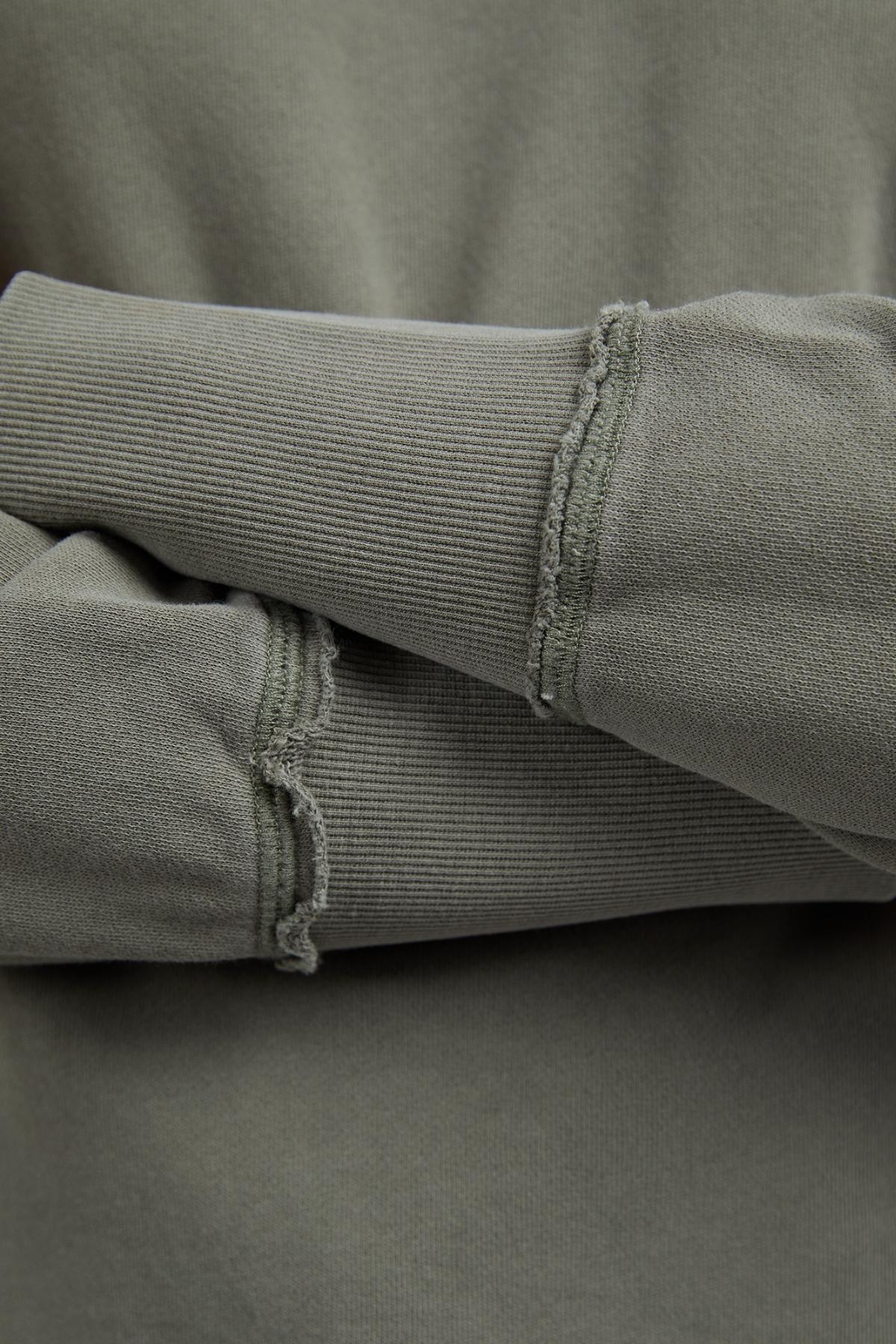 Close-up of crossed arms wearing the Foxwood Delilah Crew—an olive-green cotton jumper with a crew neck and visible unfinished seam details at the cuffs.
