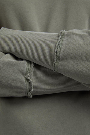 Close-up of crossed arms wearing the Foxwood Delilah Crew—an olive-green cotton jumper with a crew neck and visible unfinished seam details at the cuffs.