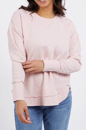 A woman wearing the light pink Foxwood Delilah Crew and blue jeans stands against a white background, smiling with one arm gently holding her opposite sleeve.