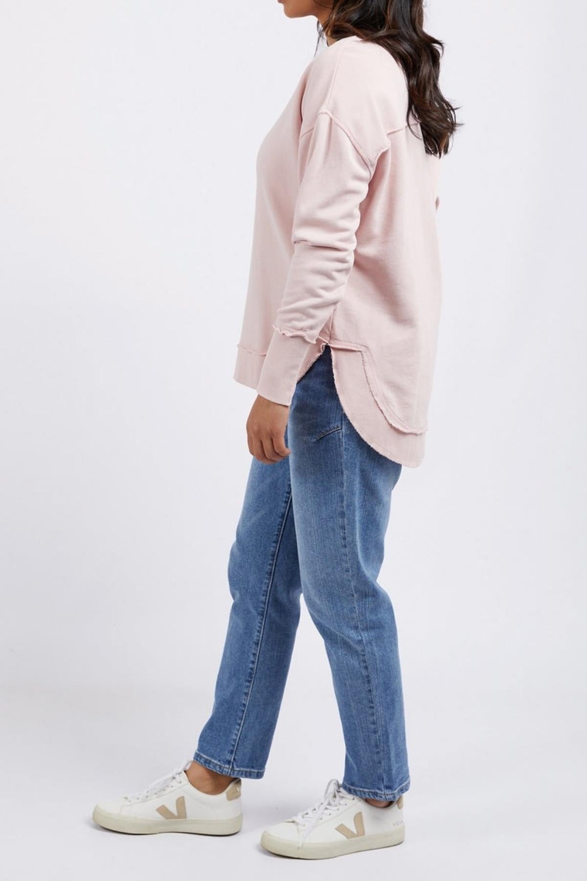 A person standing sideways wearing a light pink Foxwood Delilah Crew, blue jeans, and white sneakers with beige accents against a plain white background.