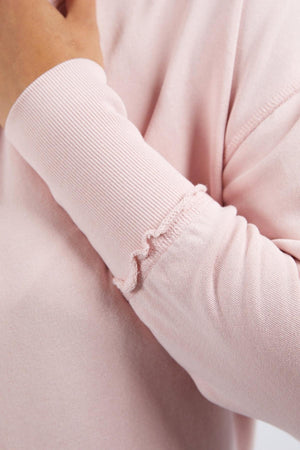 A close-up of a person wearing the light pink Foxwood Delilah Crew cotton jumper, which features ruffled sleeve edges. Their left hand is gently touching their neck near the collar.