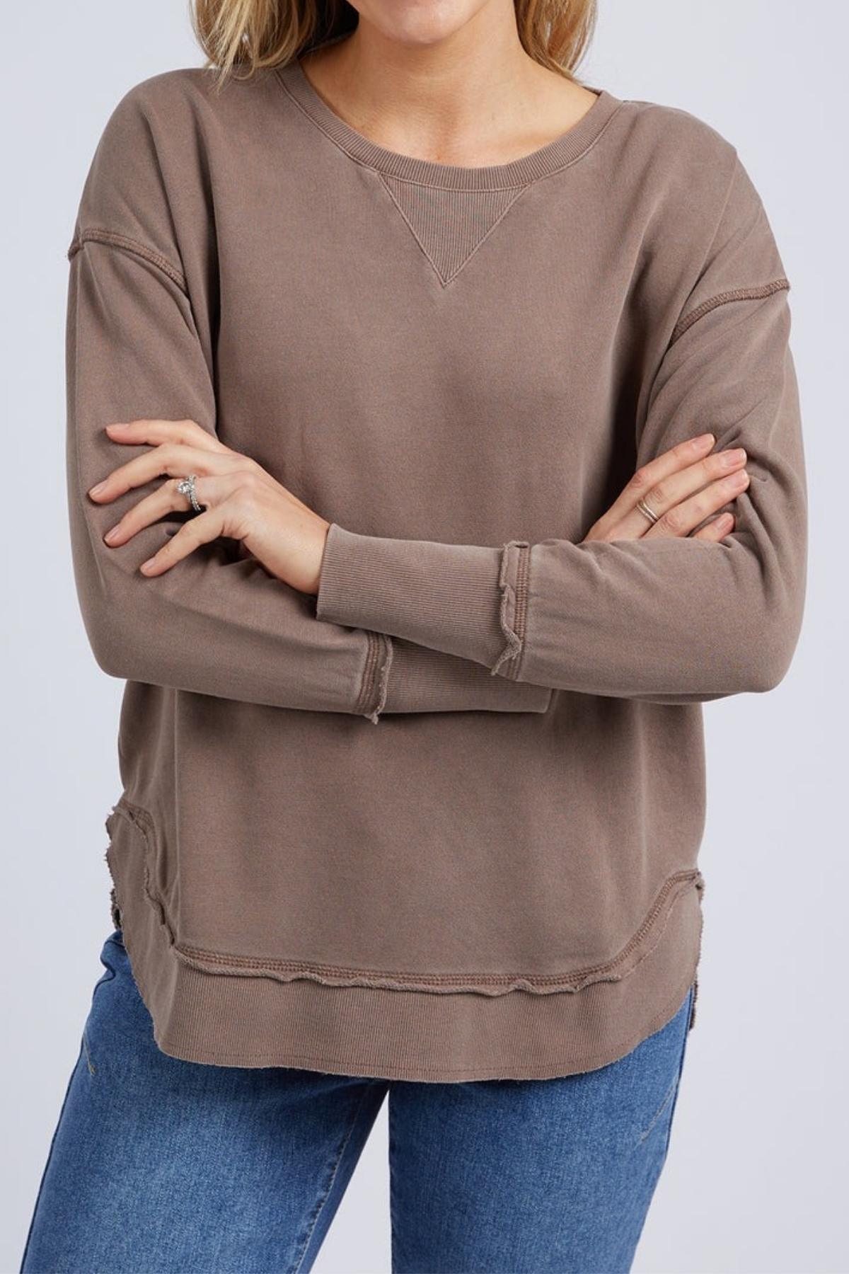 A woman stands with her arms crossed, wearing the Foxwood Delilah Crew—a loose brown cotton jumper with a crew neck and exposed seams—paired with blue jeans.