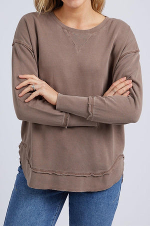 A woman stands with her arms crossed, wearing the Foxwood Delilah Crew—a loose brown cotton jumper with a crew neck and exposed seams—paired with blue jeans.