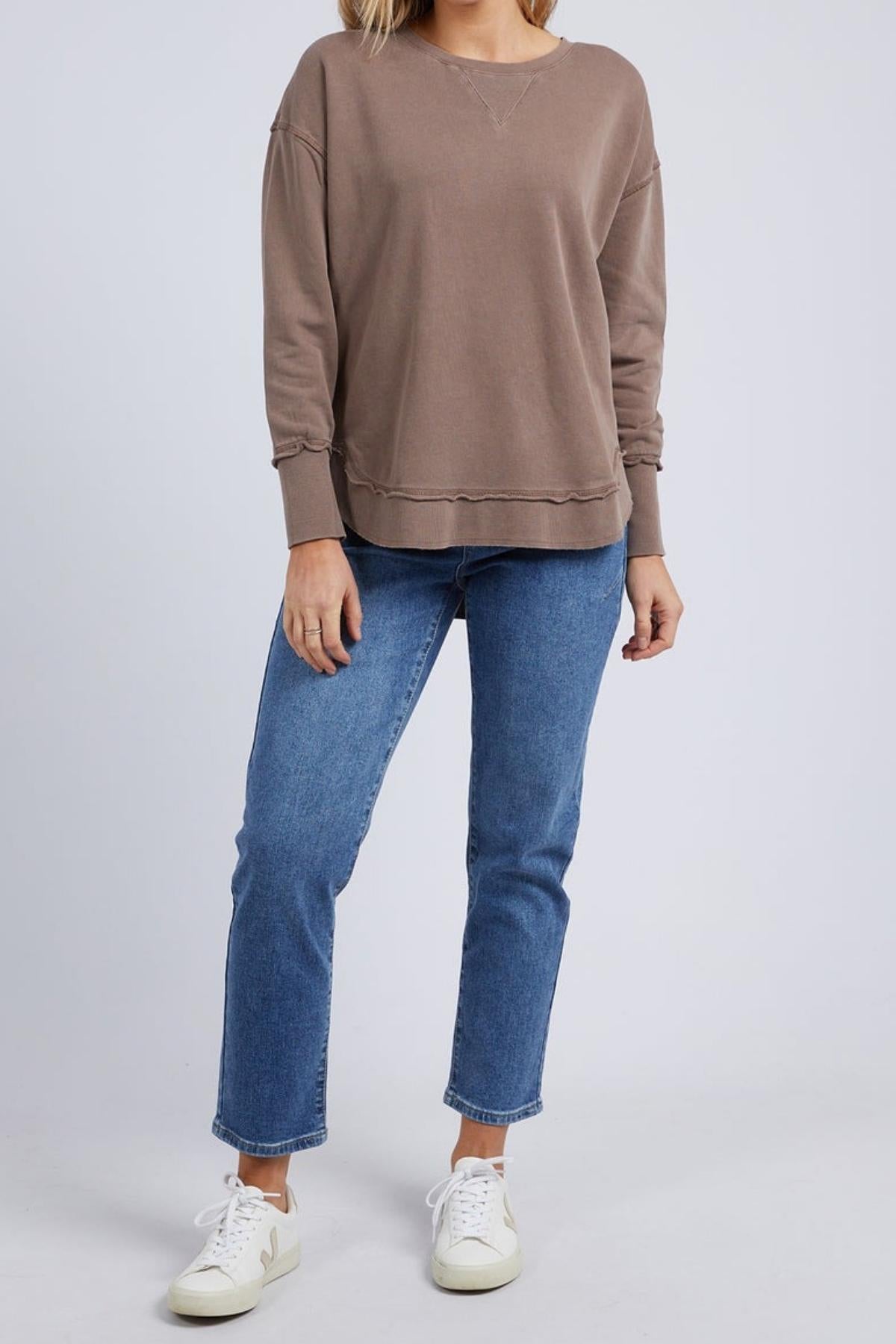 A person stands against a plain background, wearing the Foxwood Delilah Crew jumper in brown, blue jeans, and white sneakers. Their left arm rests by their side and their right hand is partly in their pocket. Their head is not visible.