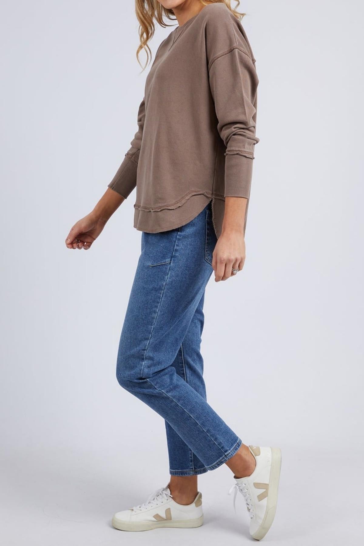 A woman stands sideways against a plain light background, wearing the Foxwood Delilah Crew in taupe, paired with blue jeans and white sneakers with beige accents. Her hair is styled in loose waves.