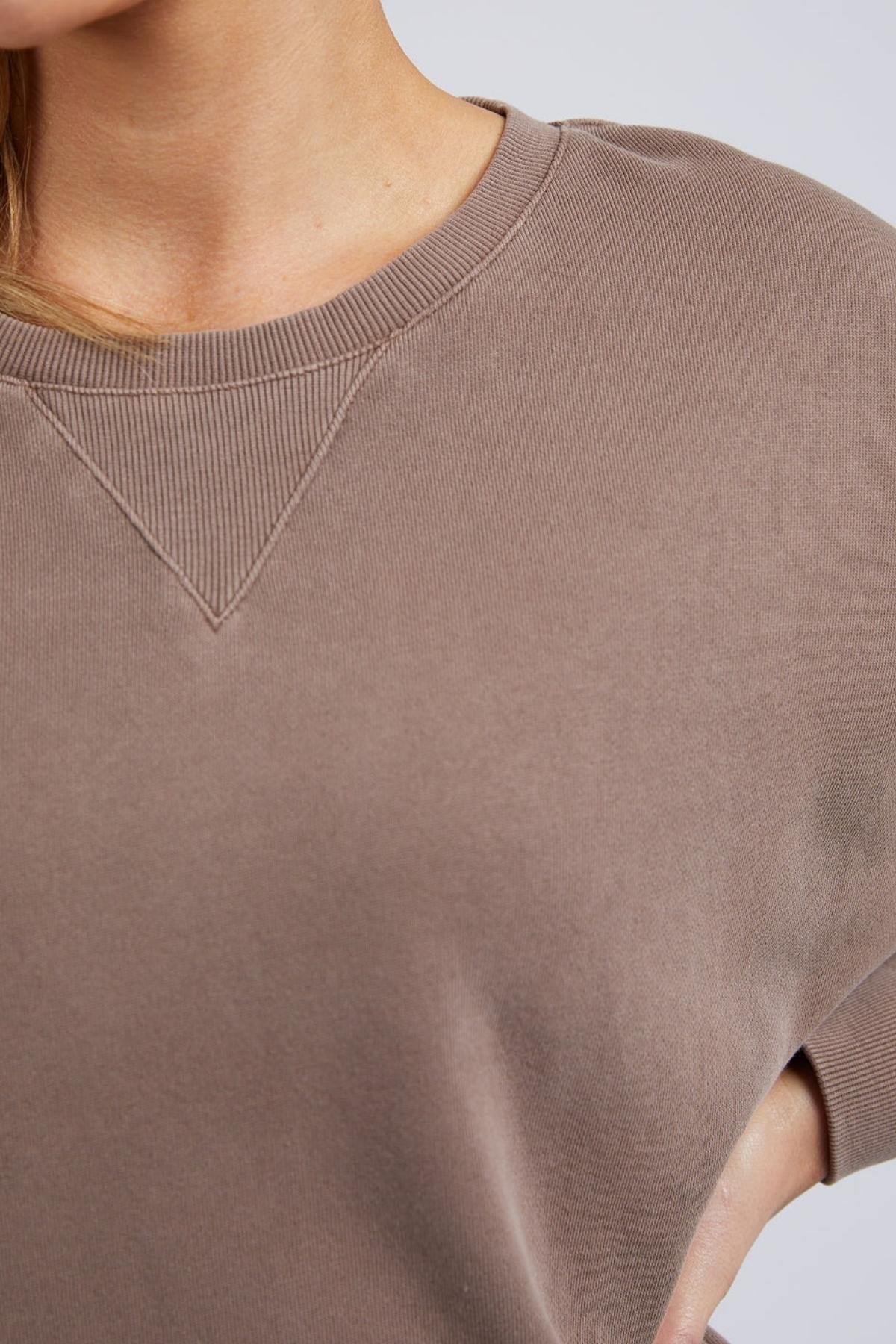 Close-up of a person wearing the Foxwood Delilah Crew in taupe cotton, featuring a ribbed crew neck. Only the neck, shoulder, and upper chest are shown against a plain background.