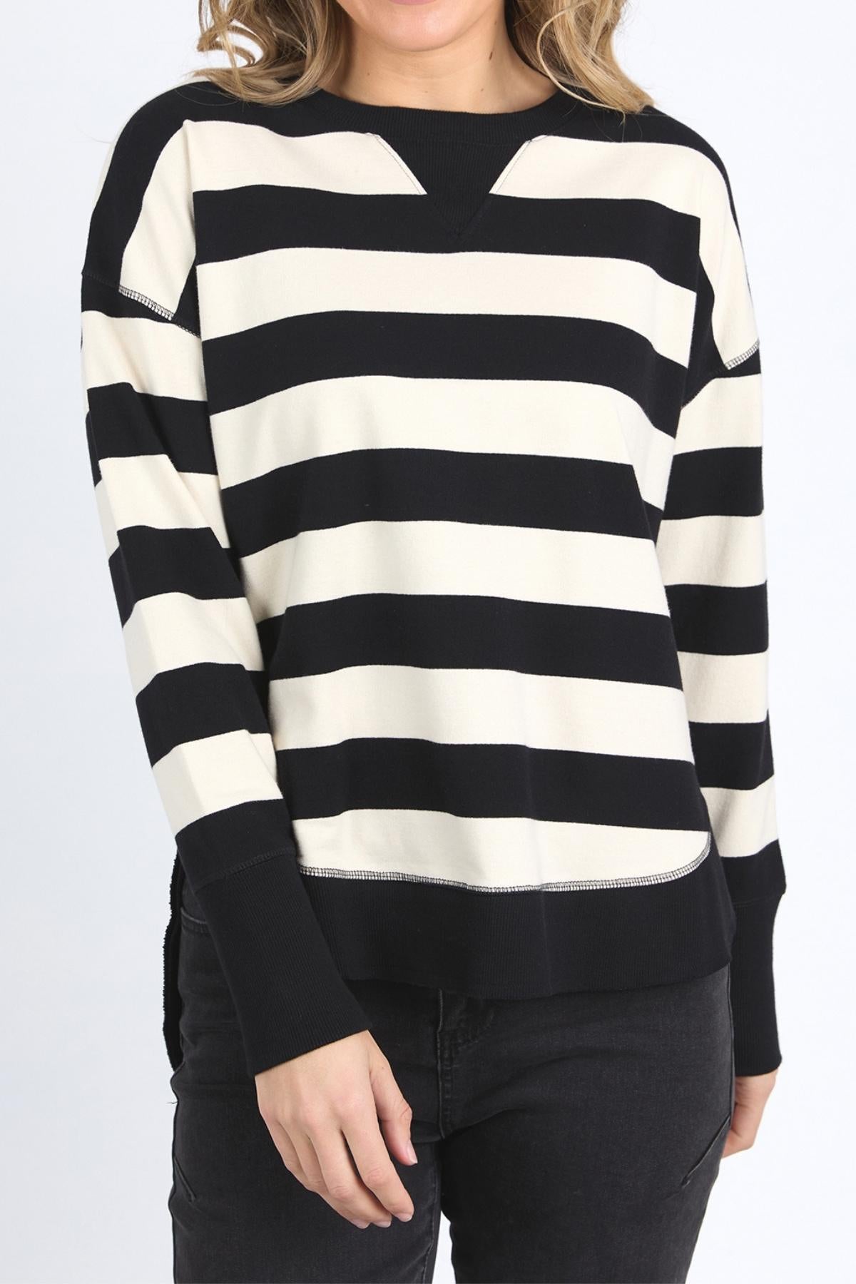 A person wearing the Foxwood Delilah Stripe Crew, a black and white striped 100% cotton jumper, with black pants stands against a plain light background. Their face is partially out of frame.