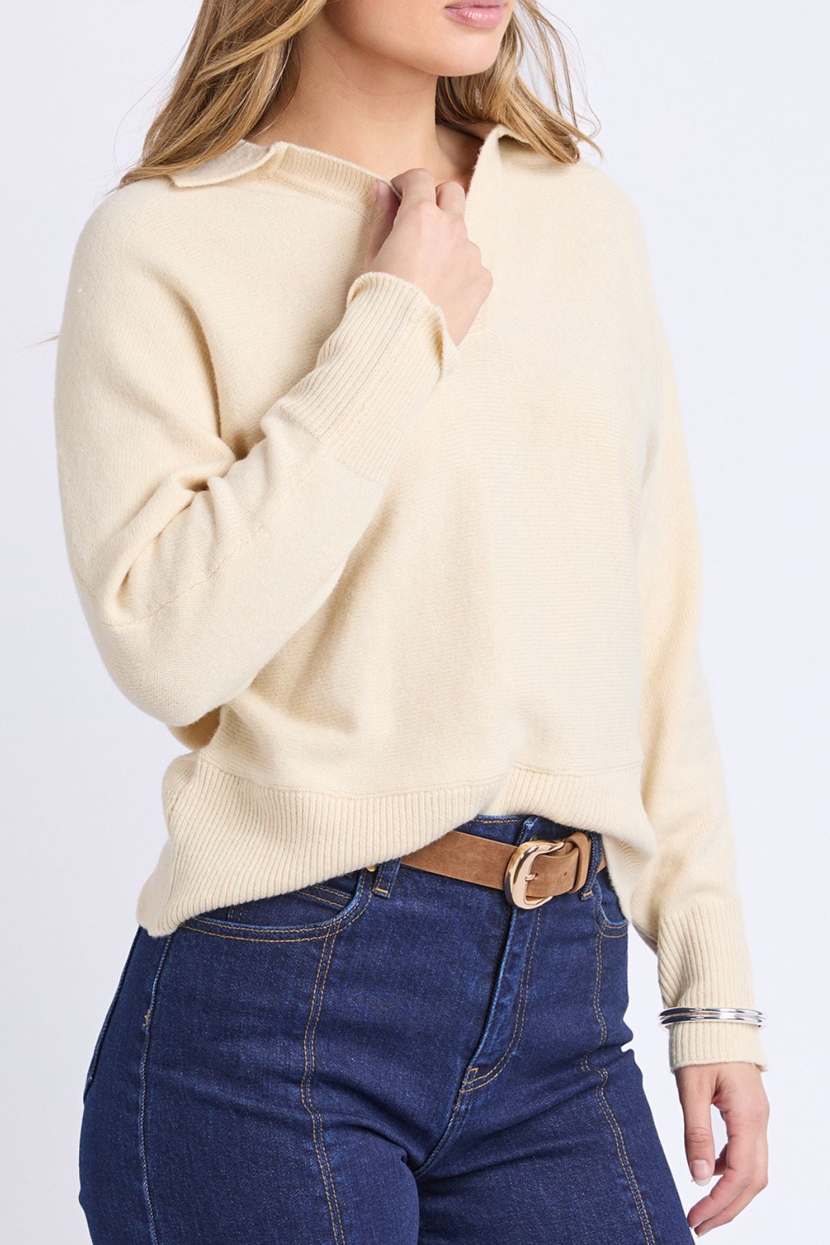 A woman with light brown hair, face partially out of frame, wears the Foxwood Elea Knit Polo in light beige and blue high-waisted jeans with a brown belt, holding the sweater near the collar.