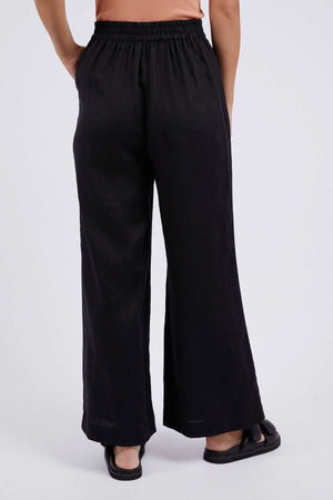 A person stands facing away, wearing relaxed fit black Foxwood Elina Wide Leg Pant in linen with an elastic waistband and wide-leg style, paired with black sandals and a peach top. The plain background is light-colored.