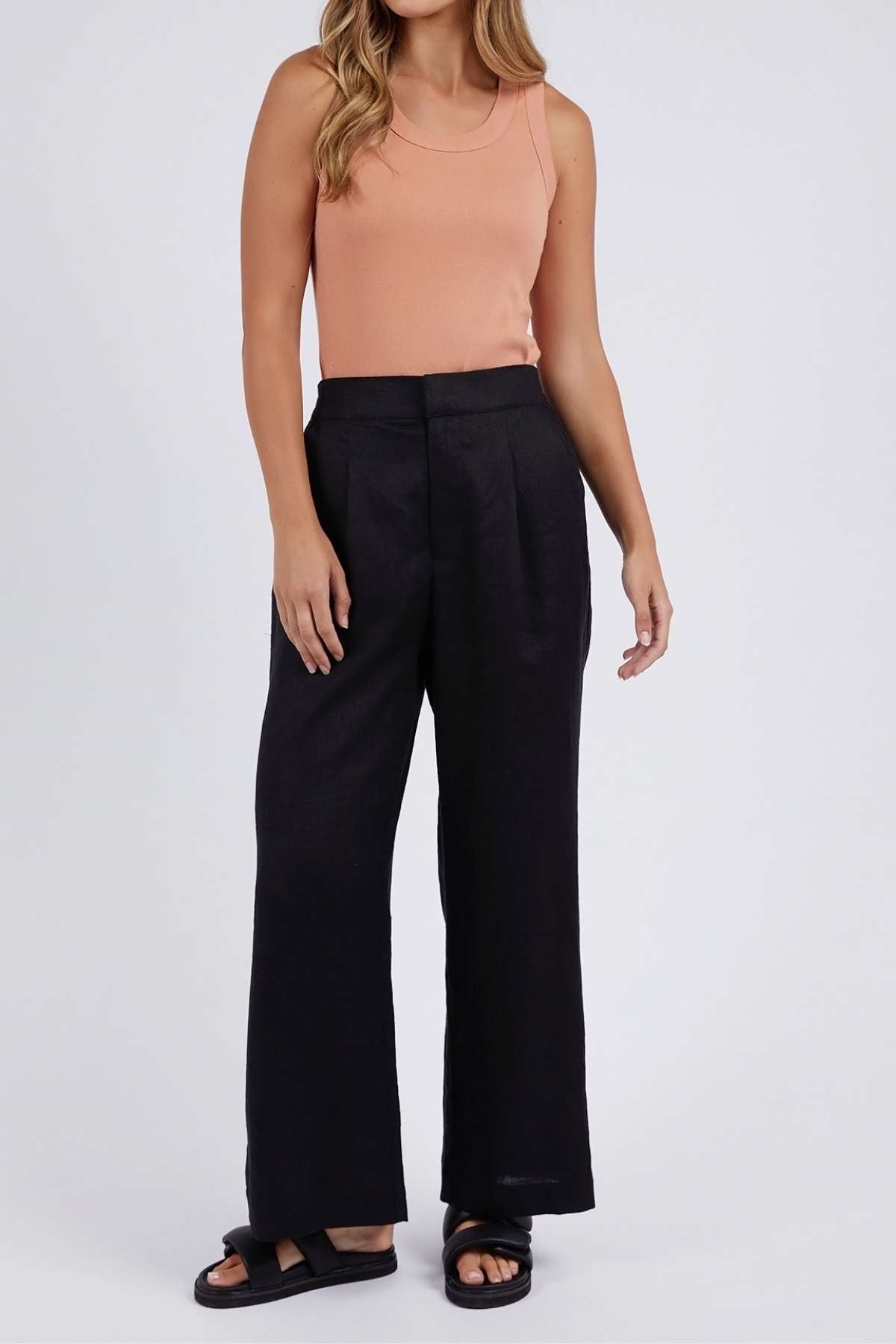 A person wearing a sleeveless peach top, the relaxed fit high-waisted Foxwood Elina Wide Leg Pant in black linen, and black sandals stands against a plain light background.