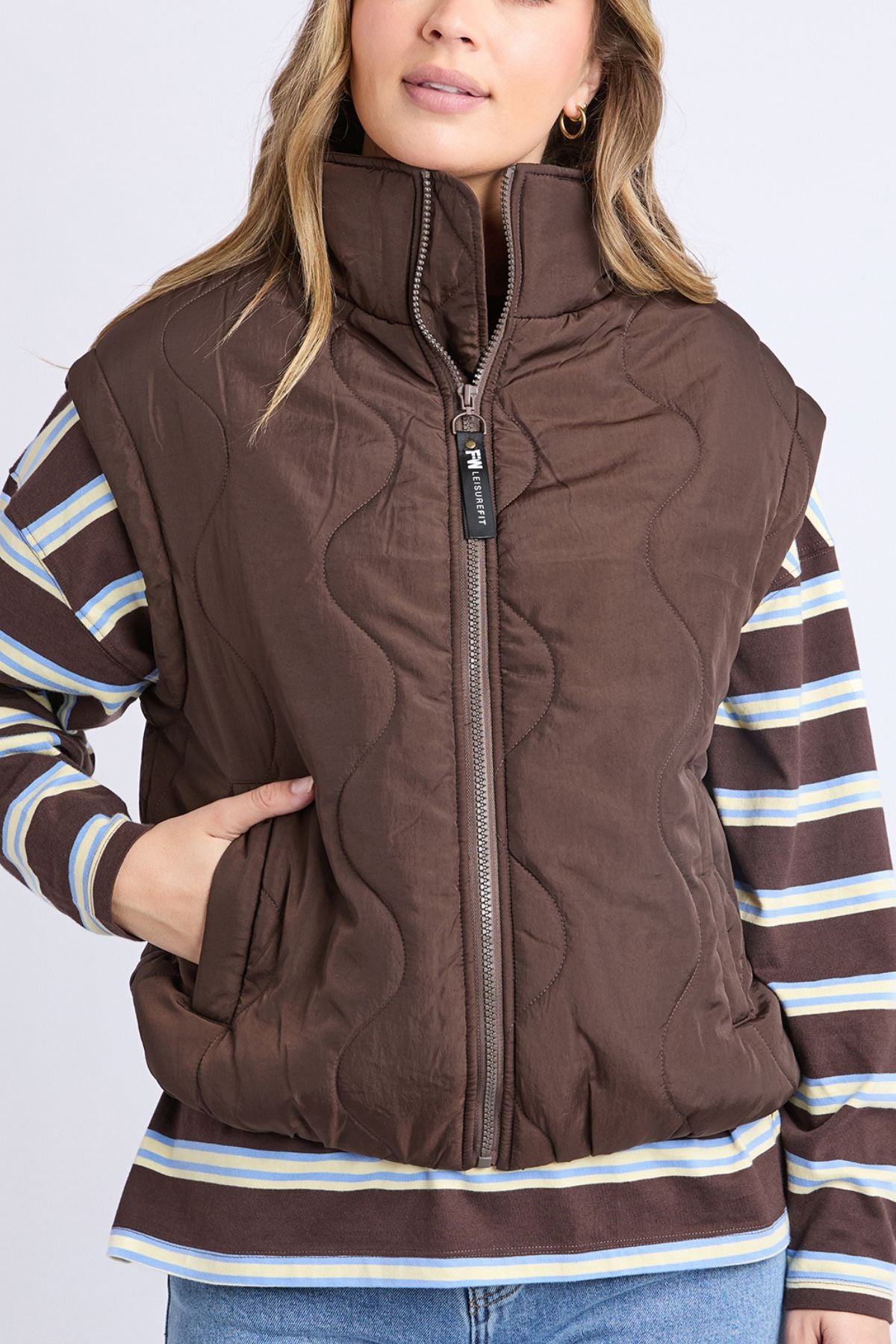 A woman models the Foxwood Elite Vest, a brown quilted zip-up with a high collar, paired over a striped long sleeve and her left hand in the pocket, standing before a plain light background.