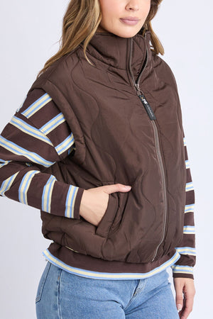 A woman wears the Foxwood Elite Vest, a brown quilted zip-up, over a striped long-sleeve shirt and light blue jeans. She has one hand in her vest pocket, with her face partially visible.