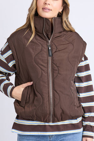 A woman models the Foxwood Elite Vest, a brown quilted zip-up with a high collar, paired over a striped long sleeve and her left hand in the pocket, standing before a plain light background.