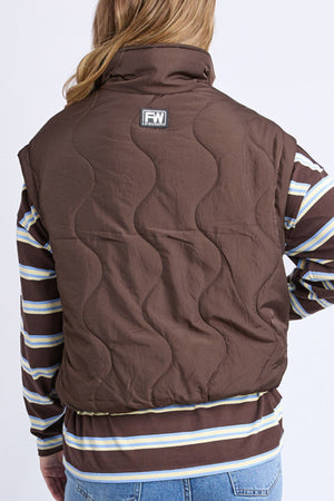 A person with long hair is seen from behind wearing the Foxwood Elite Vest over a brown, blue, and white striped long-sleeve shirt and light blue jeans.