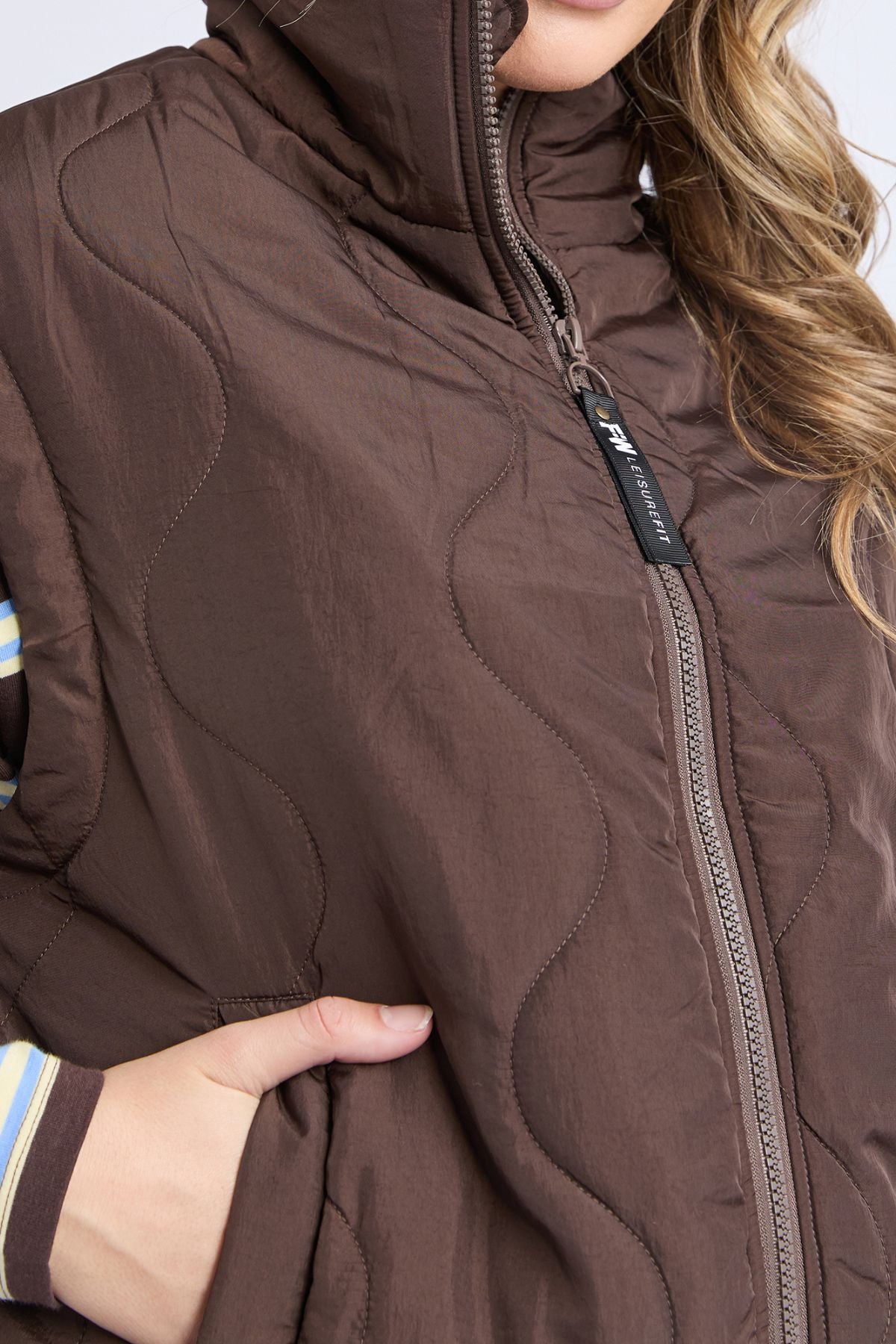 Someone wears the Foxwood Elite Vest—a brown, quilted vest with a high collar and zipper—over a striped shirt, their hand in its pocket. Wavy hair falls to one side, highlighting the vest's practical design.