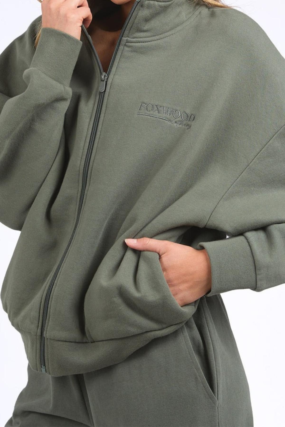 Wearing the olive green Foxwood Ember Zip Thru brushed cotton fleece jacket and matching pants, one hand in a pocket; the zip thru features a "FOXWOOD" front chest logo print.