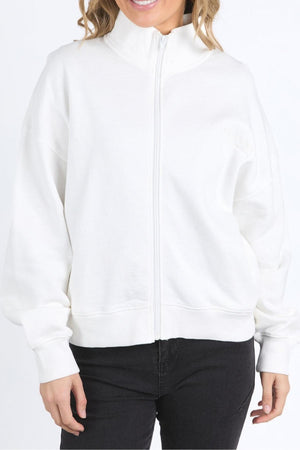 A woman in a plain white Foxwood Ember Zip Thru jacket with a high collar and subtle chest logo stands against a white background, paired with black pants. Her face is partly out of frame.