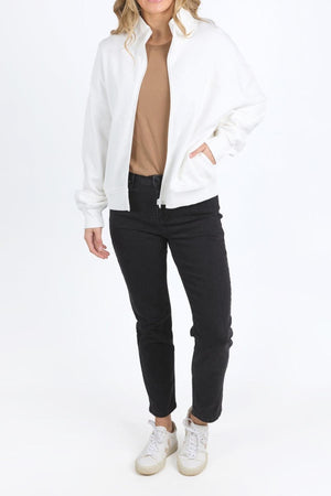 Wearing the Foxwood Ember Zip Thru brushed cotton fleece jacket with chest logo over a tan shirt, black jeans, and white sneakers, the person stands against a plain white background with hands in pockets and face partly out of frame.