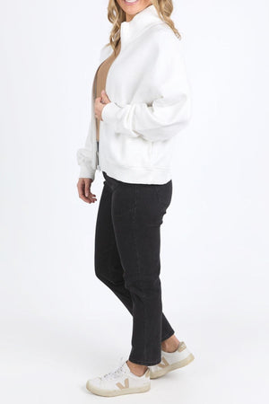 A woman stands sideways wearing the Foxwood Ember Zip Thru white brushed cotton fleece jacket with a chest logo, beige top, black jeans, and white sneakers with beige accents against a plain white background.