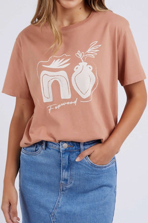 Someone wears the Foxwood Etched Geo Tee, a light brown 100% cotton crew neck with abstract white line art and "Foxwood," paired with a blue denim skirt. Their left hand rests casually in the skirt pocket.