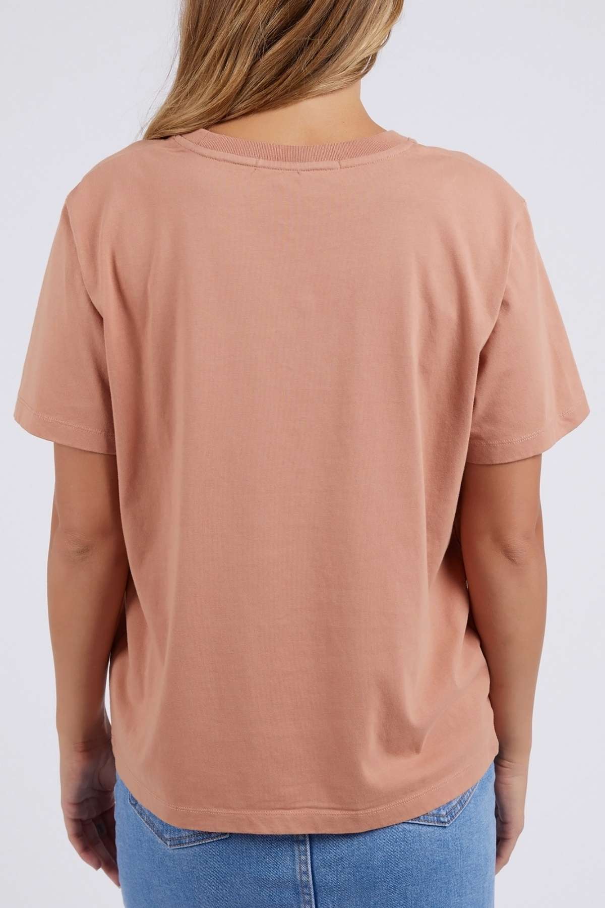 A person with long hair stands against a plain background wearing light blue jeans and the Foxwood Etched Geo Tee—a relaxed fit, peach-colored shirt made of 100% cotton.