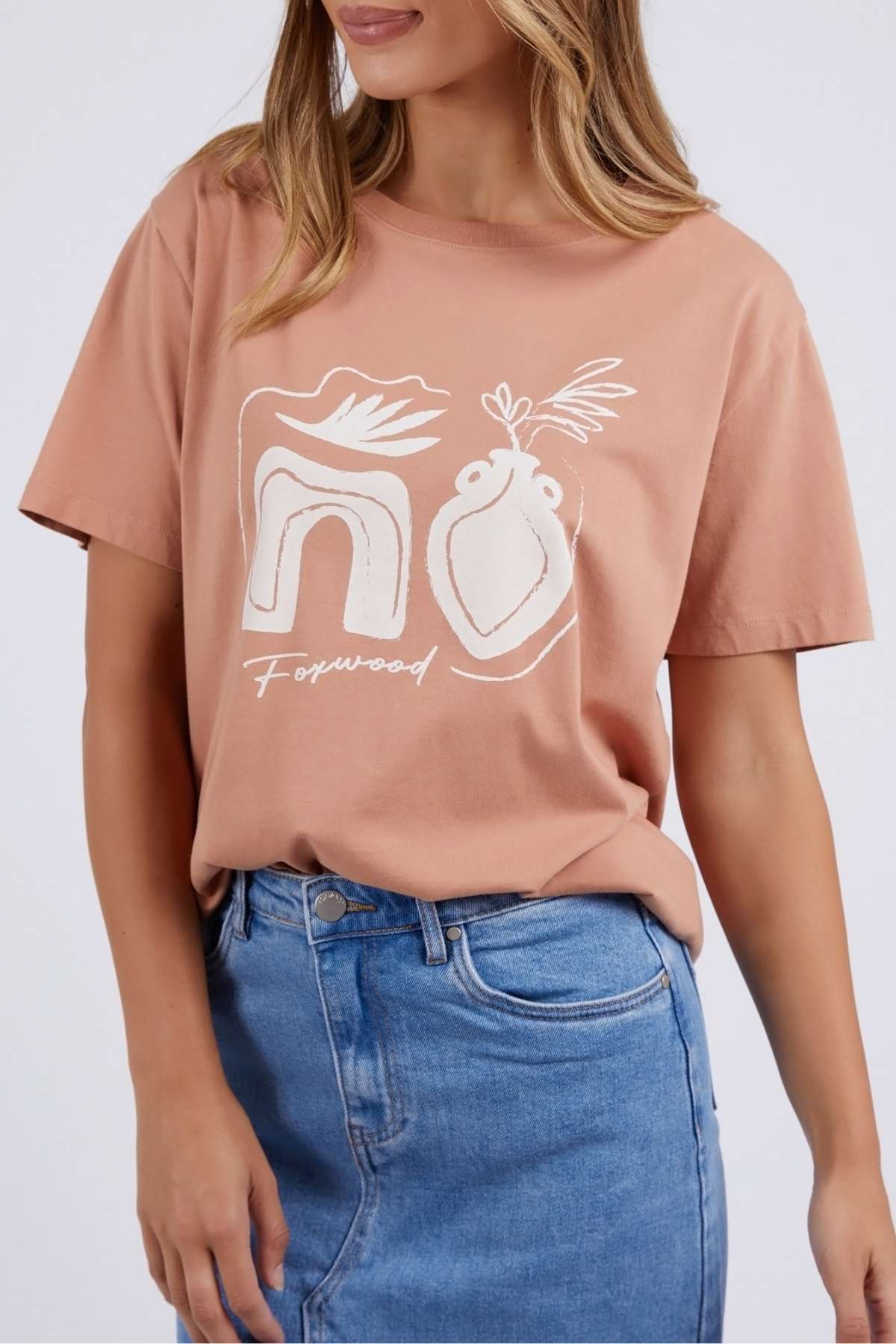 The woman is wearing a peach Foxwood Etched Geo Tee, tailored in a relaxed fit with 100% cotton. Featuring abstract white line art and "Foxwood" on the front, it's ideally paired with a blue denim skirt.