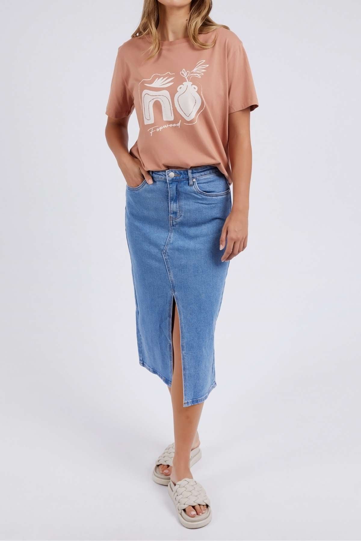 The person wears a relaxed fit brown tee similar to the Foxwood Etched Geo Tee, featuring abstract designs and "NO," paired with a blue denim midi skirt with a front slit. Light beige woven sandals complete the ensemble against a plain white background.