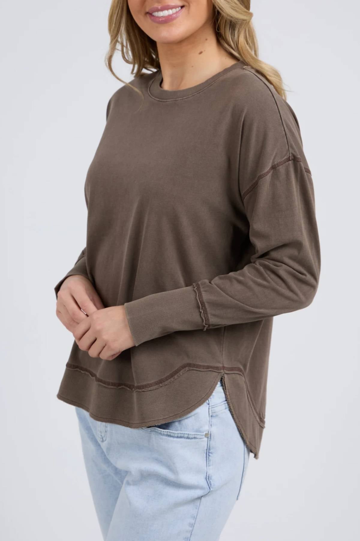 A woman with wavy blonde hair smiles, standing with hands clasped in front, wearing the Foxwood Farrah Long Sleeve relaxed fit top in brown and light blue jeans against a light gray background.