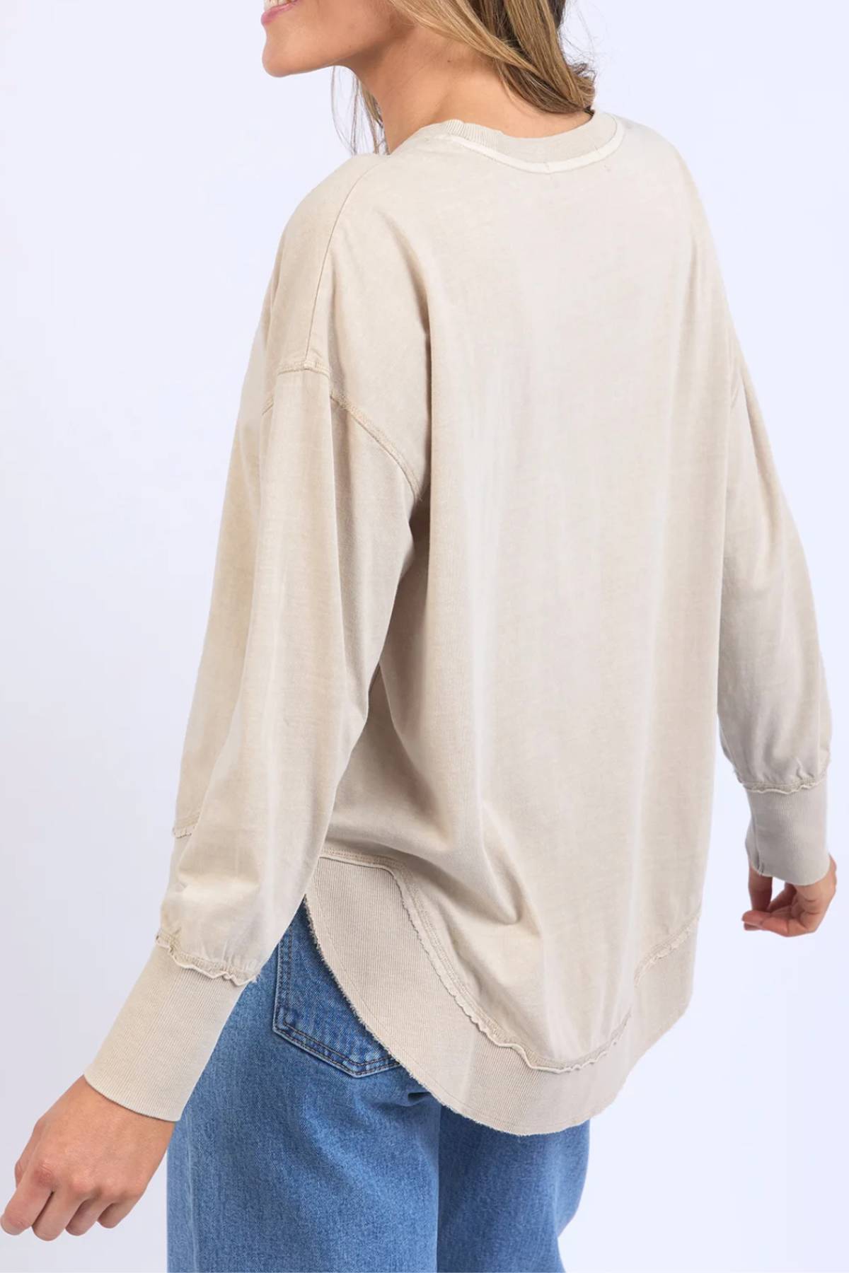 A woman, shown from the back and slightly turned to the side against a plain light background, wears the Foxwood Farrah Long Sleeve in beige with blue jeans.