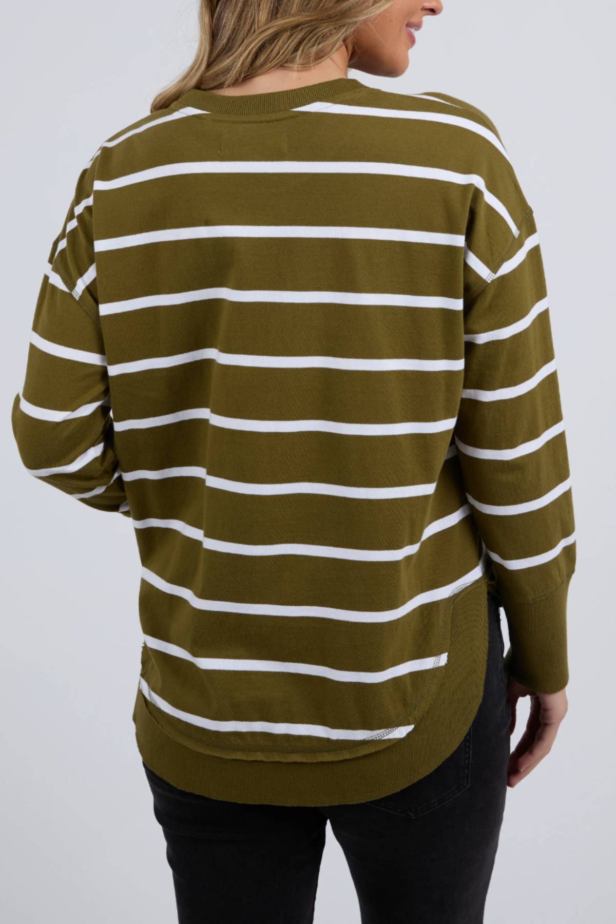 A person with light brown hair is shown from the back, wearing the Foxwood Farrah Stripe Long Sleeve jumper in olive and white, made from 100% Fresh Pima Cotton, featuring a rounded hem, long sleeves, and paired with black pants.