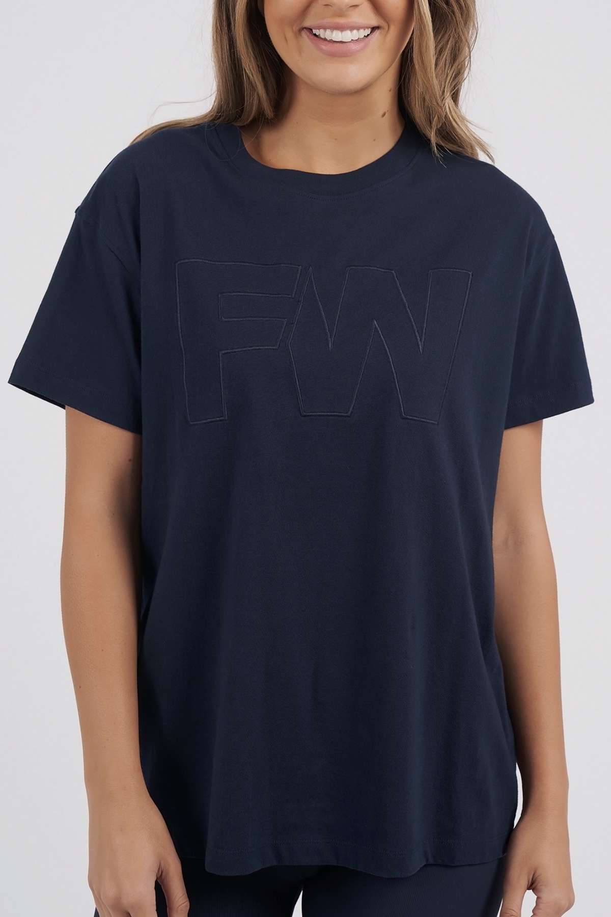 A woman smiles against a plain background wearing the Foxwood FW Embroidery Tee, a loose-fit 100% cotton top featuring subtle satin-embroidered "FW" detailing on the front for an elegant touch.