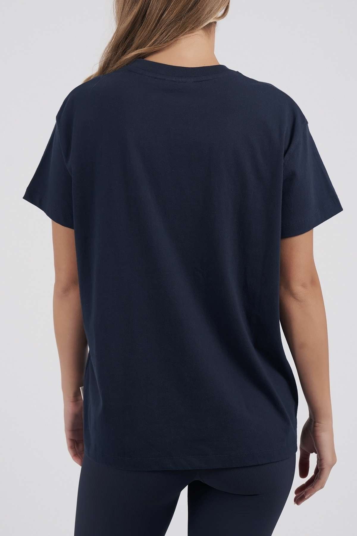 A woman with long hair stands with her back to the camera, wearing a plain navy Foxwood FW Embroidery Tee and dark pants against a light, neutral background.