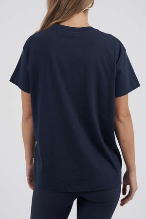 A woman with long hair stands with her back to the camera, wearing a plain navy Foxwood FW Embroidery Tee and dark pants against a light, neutral background.